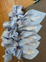 Set of light blue scrunchies with personalized names on a wooden surface.