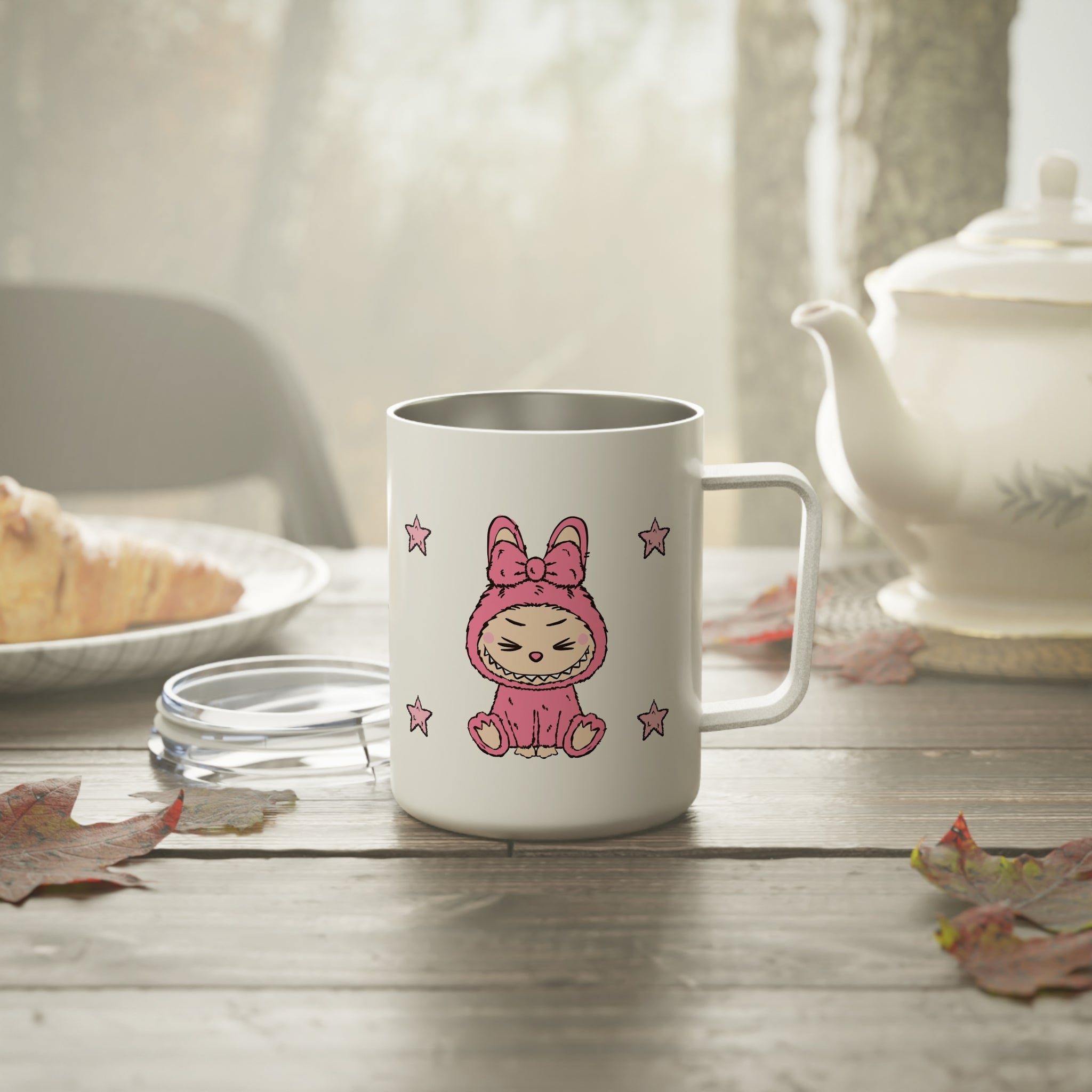 White mug with a pink Labubu monster on a wooden table with a teapot and bread in the background.